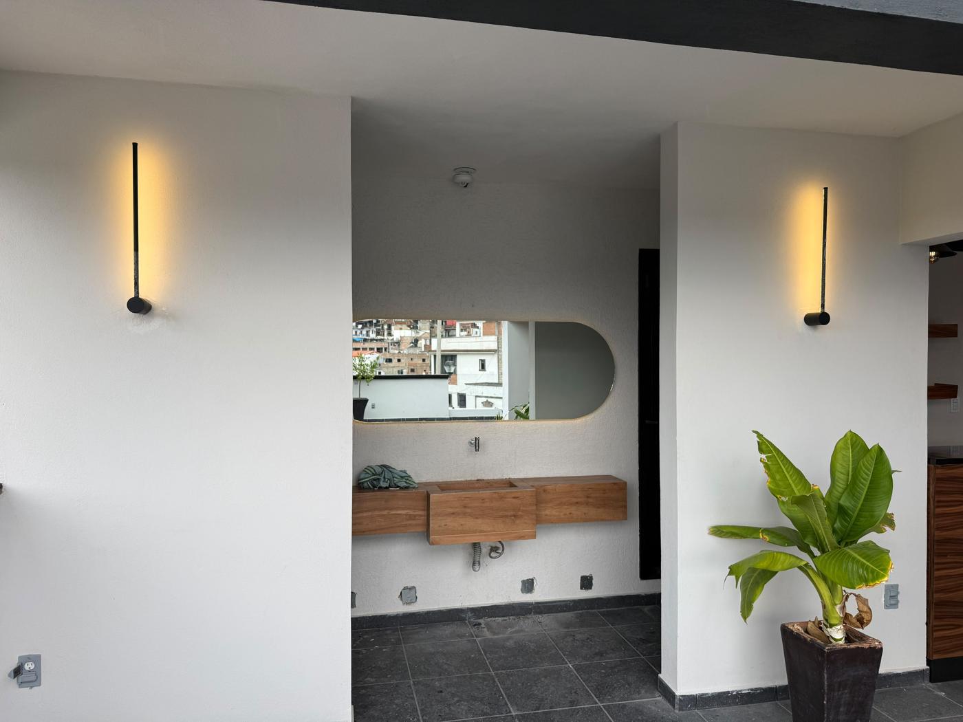 Muro con dos luminarias lineales cálidas, espejo con luz perimetral y lavabo de madera